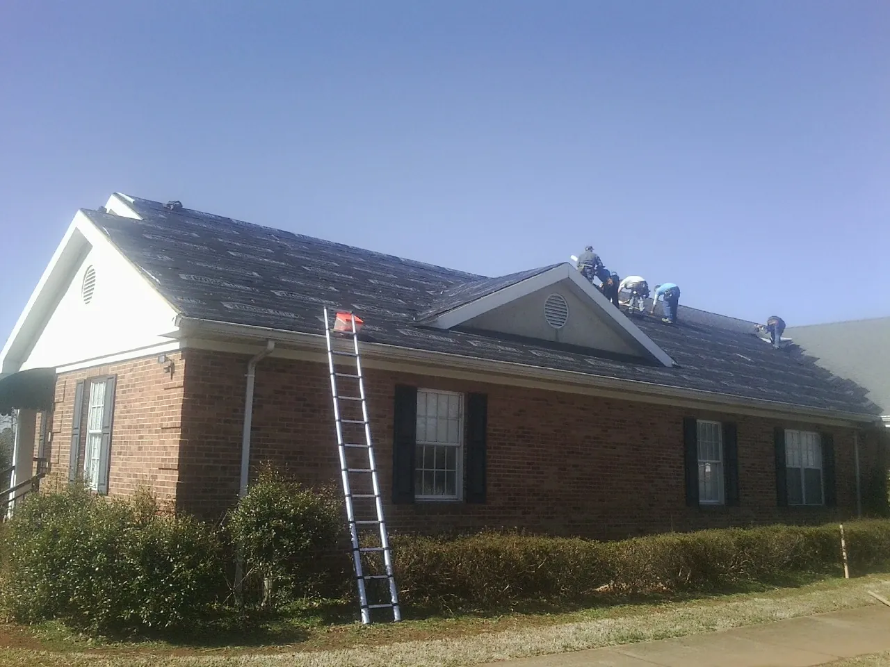 Crew tearing shingles off a brick ranch roof with a ladder set — Emergency Roof Repair work by our crew in Holdingford