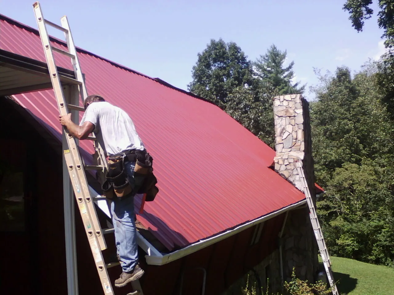 Roofer on a ladder beside a red metal roof and stone chimney — Soffit & Fascia Repair by Roofing Brothers in Holdingford